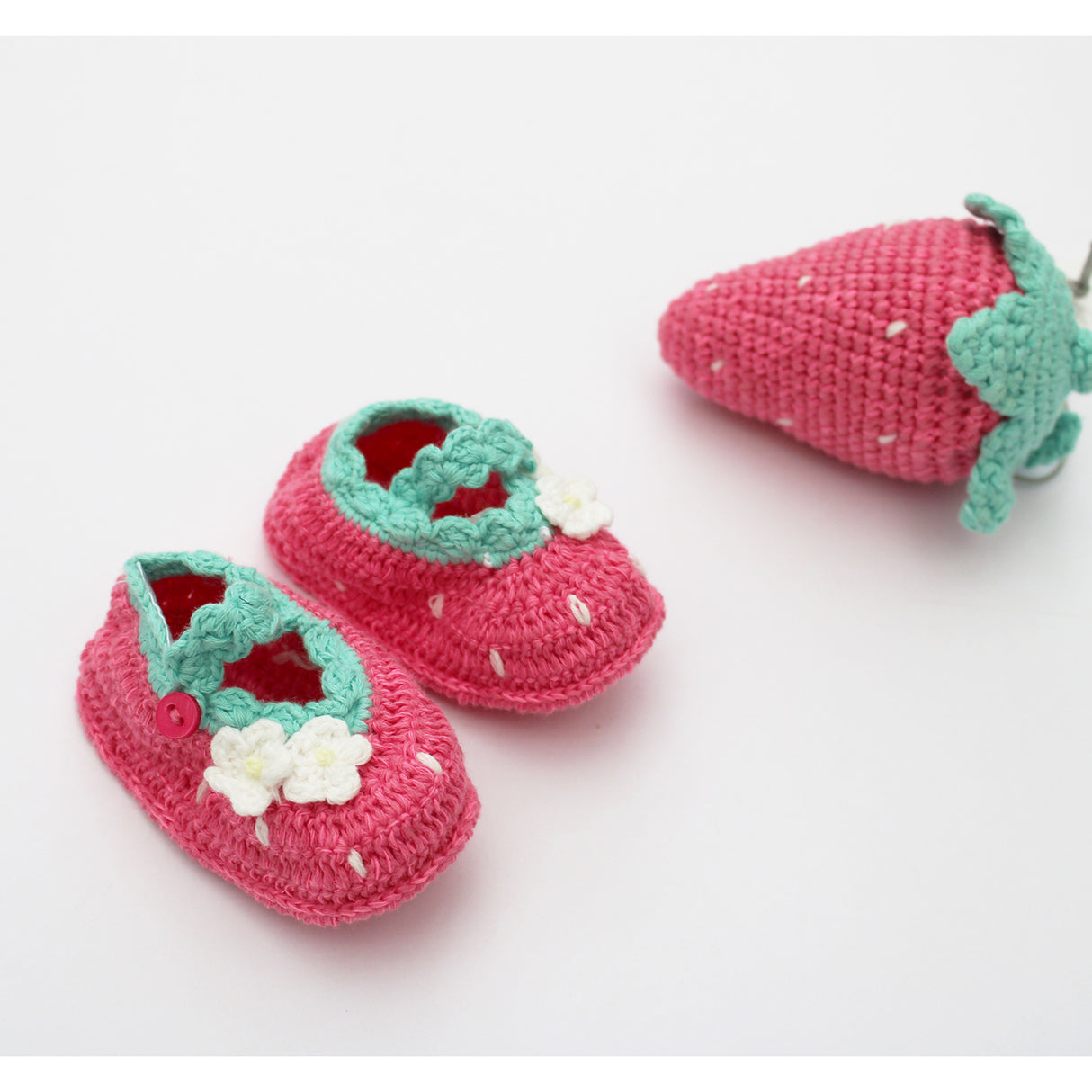 Pair of pink strawberry crochet baby shoes with green straps and white flowers on a light background