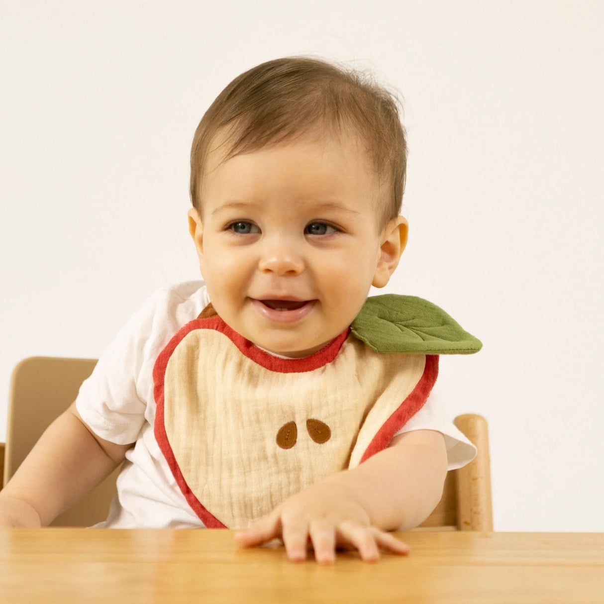 Smiling baby wearing the Oli & Carol Pepa The Apple Bib, seated at a wooden table