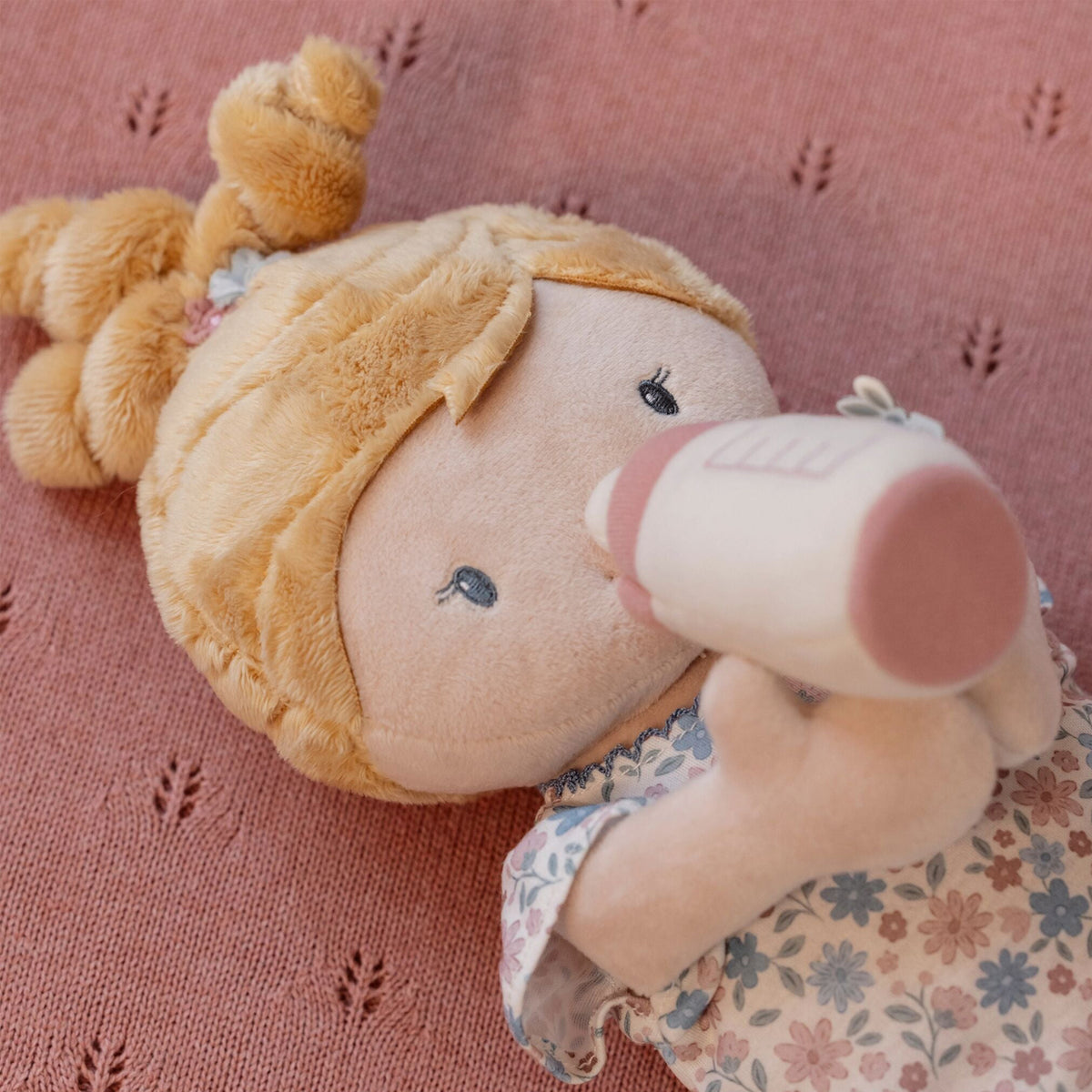 A close-up of soft baby doll Julia with blonde hair and a floral romper, holding a pink and white bottle against a pink textured knit background.
