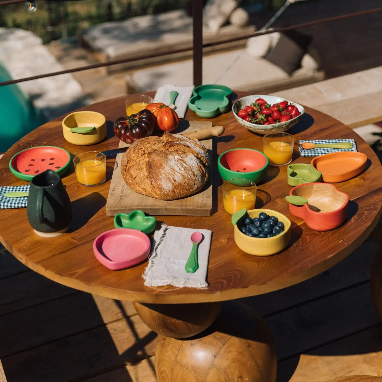 Wooden table with colourful childrens wooden plates, bowls, and food items outdoors.