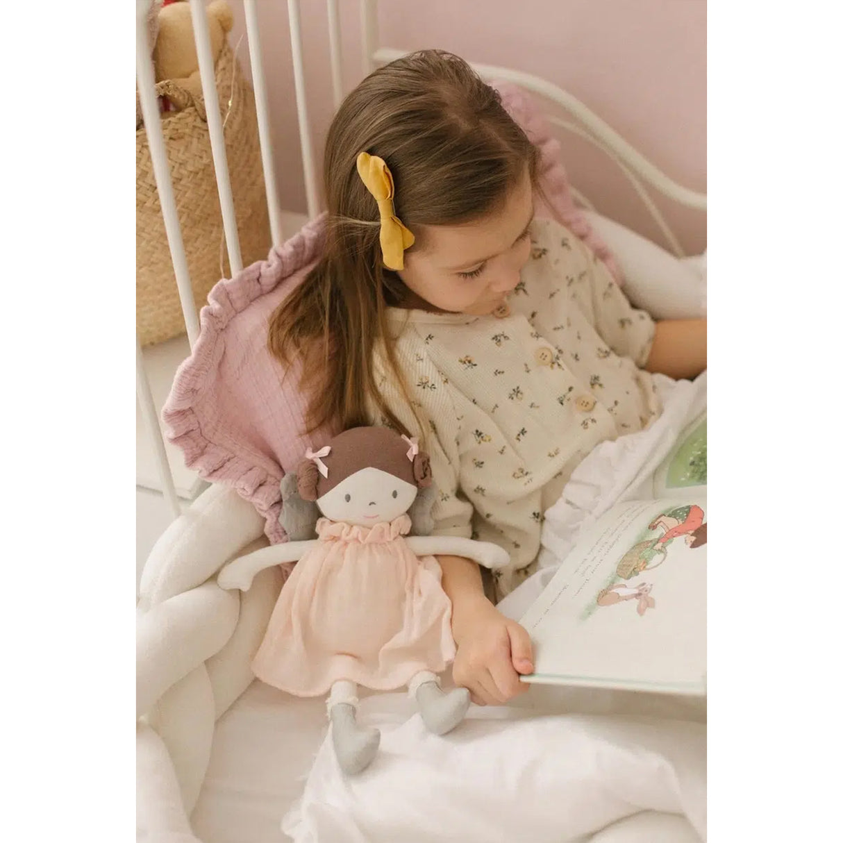 Child reading a book with a doll wearing a pink dress and brown hair and grey fairy wings and feet in a crib