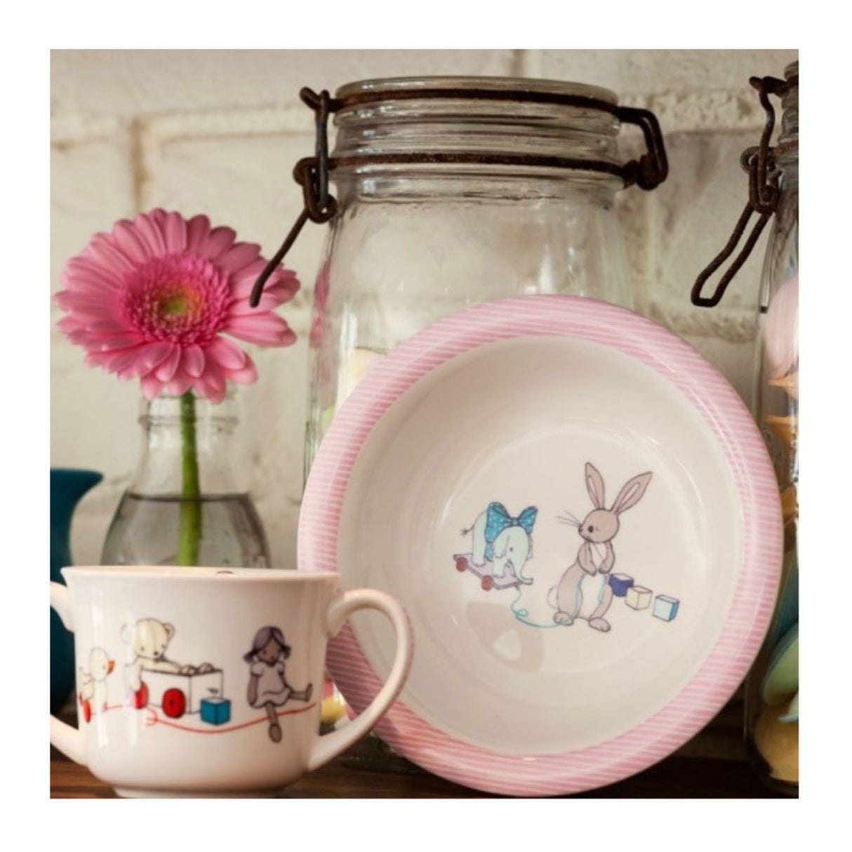 Ceramic mug and plate with cartoon designs on a wooden surface, featuring a pink flower and glass jar in the background.