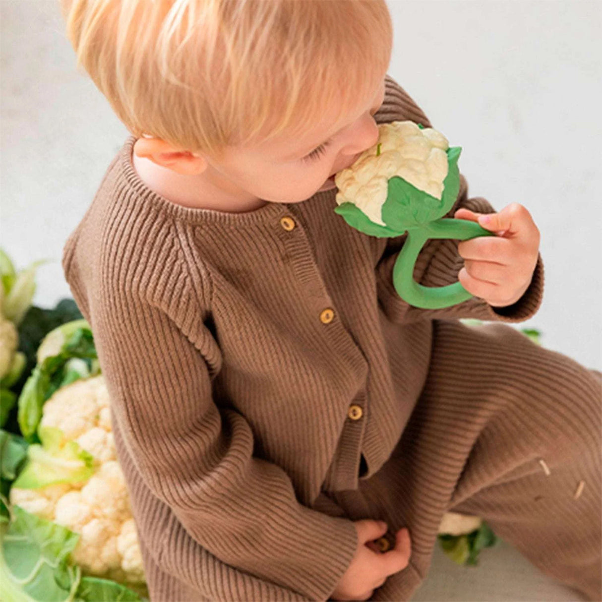 Child in a brown outfit holding a green natural rubber teething ring in the shape of a cauliflower sitting among real cauliflowers.