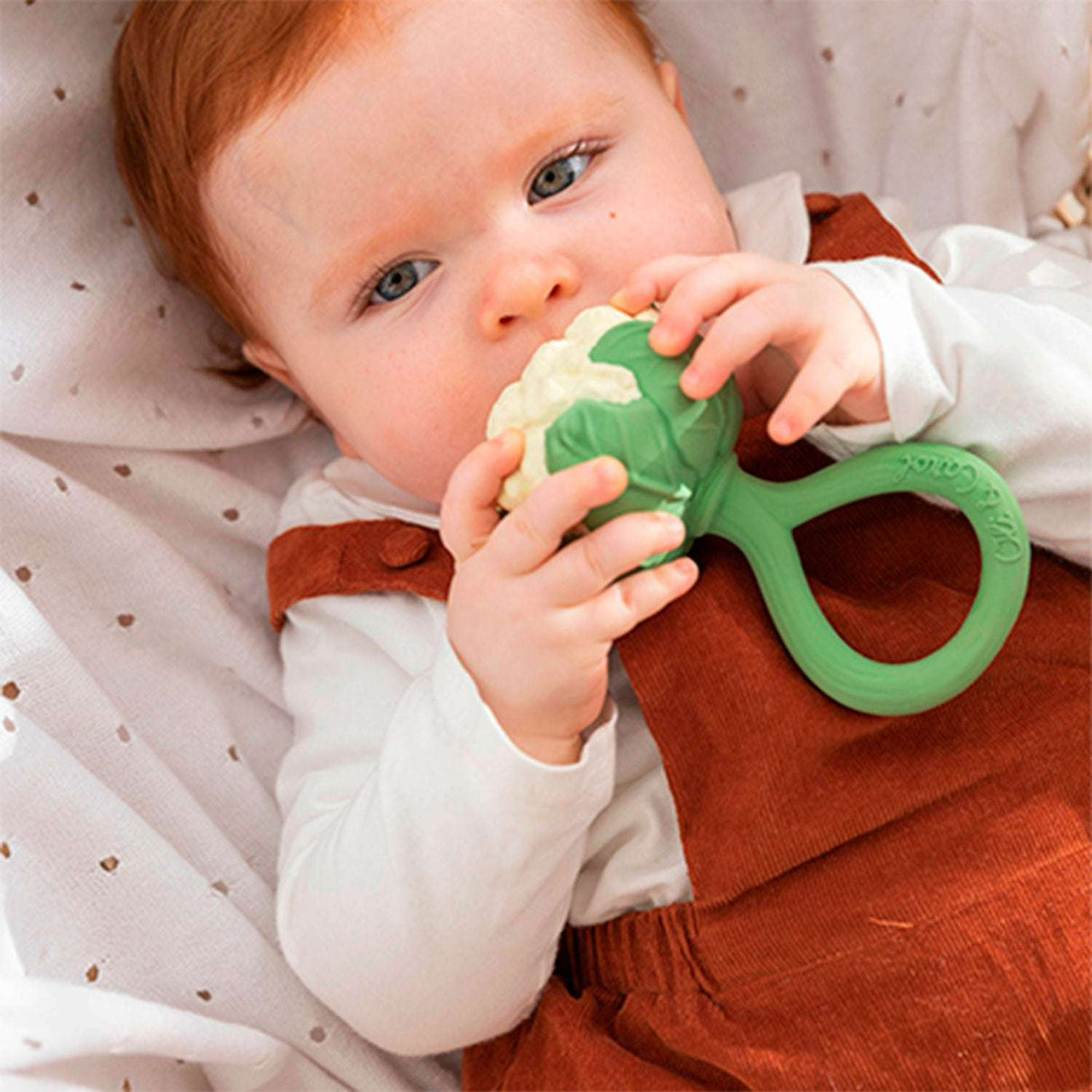 Baby holding a green natural rubber teething ring in the shape of a cauliflower with a soft bedding background