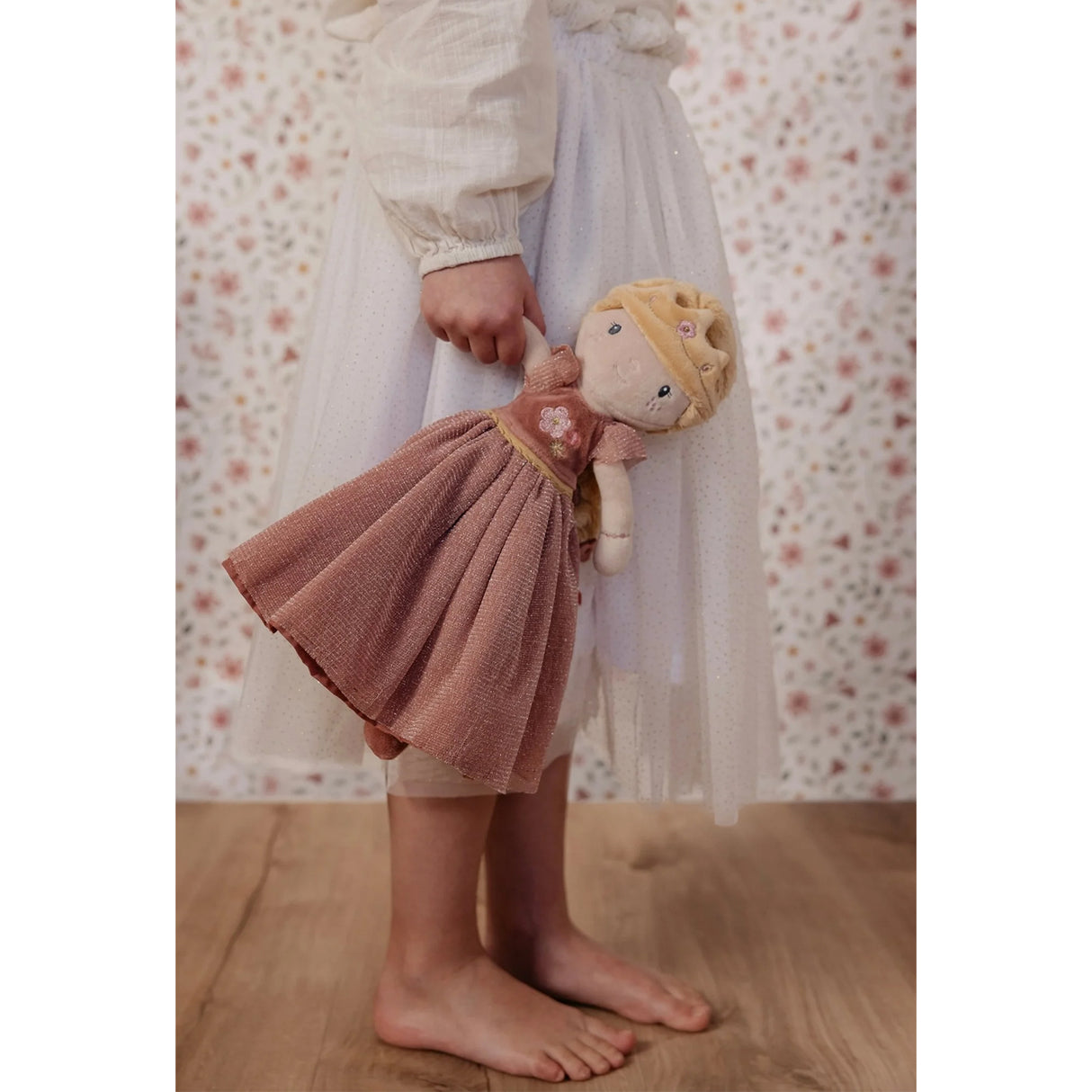 Child holding a doll wearing a pink gown with floral pattern and floral wall in the background
