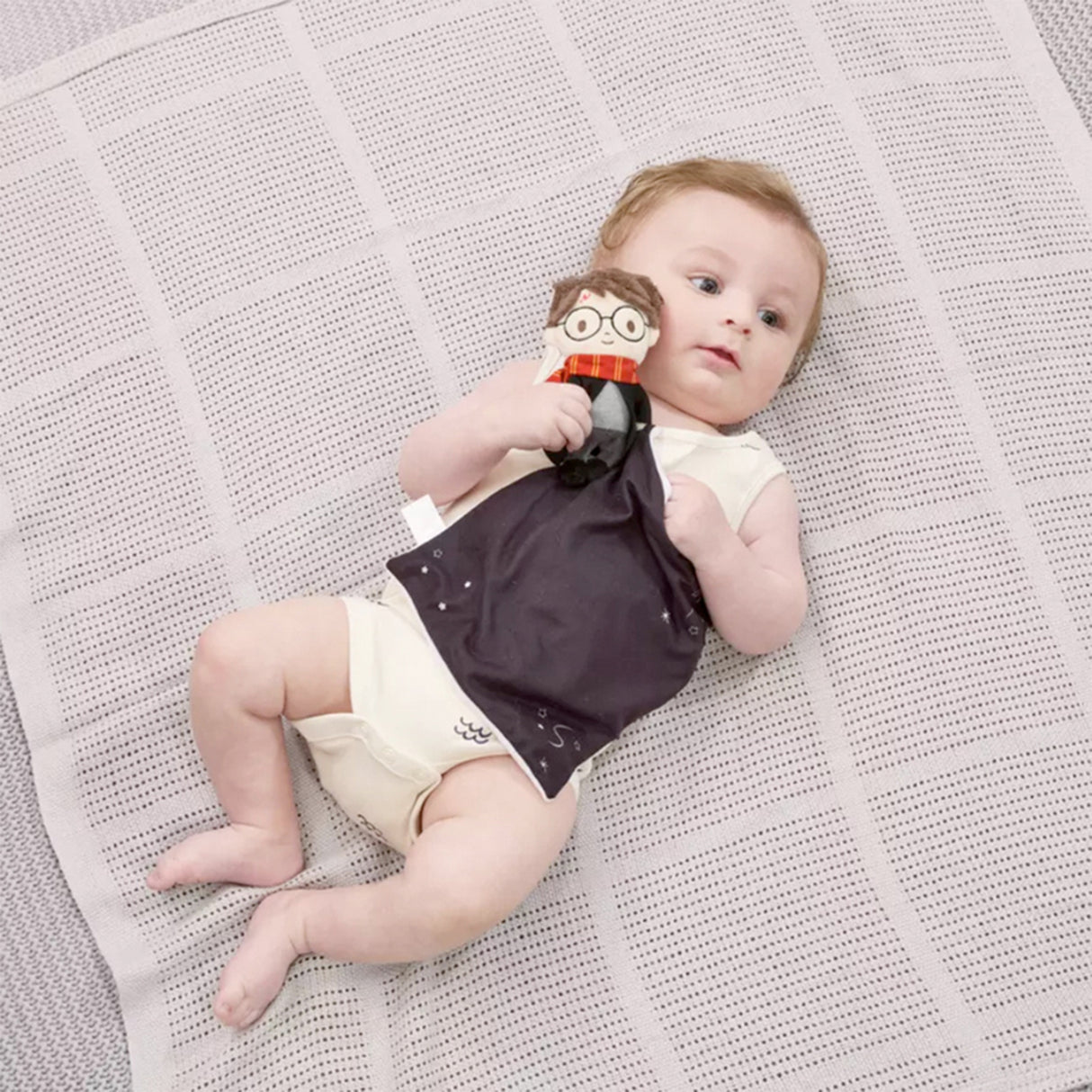 Baby lying on a textured blanket holding a Harry Potter themed comfort blanket with a plush toy of Harry Potter in his signature school robes and Gryffindor scarf attached to the corner.