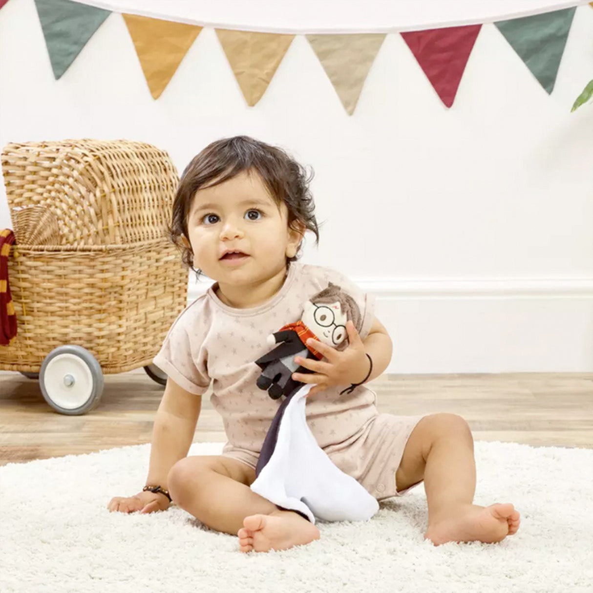 Child sitting on a white rug holding a tHarry Potter themed comfort blanket with a plush toy of Harry Potter in his signature school robes and Gryffindor scarf attached to the corner, with a colorful banner in the background