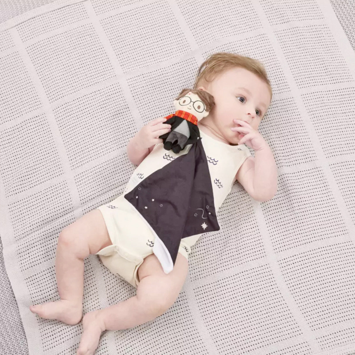 Baby lying on a textured blanket wearing a onesie holding a Harry Potter themed comfort blanket with a plush toy of Harry Potter in his signature school robes and Gryffindor scarf attached to the corner.