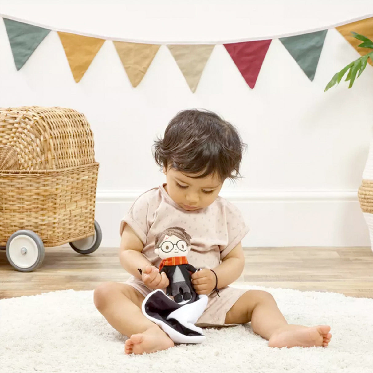 Child sitting on a white rug holding a Harry Potter themed comfort blanket with a plush toy of Harry Potter in his signature school robes and Gryffindor scarf attached to the corner, a colorful banner in the background