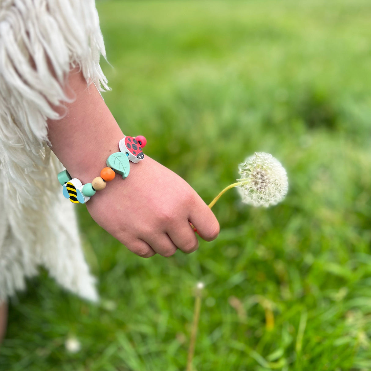 Child's hand holding a dandelion with a colorful beaded bracelet on a grassy background