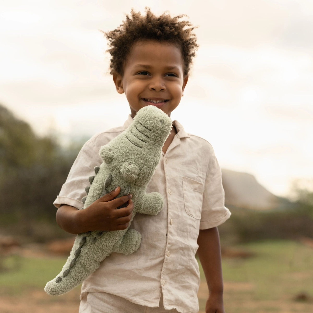 A young boy holding a soft crocodile toy with a soft-focus natural background.