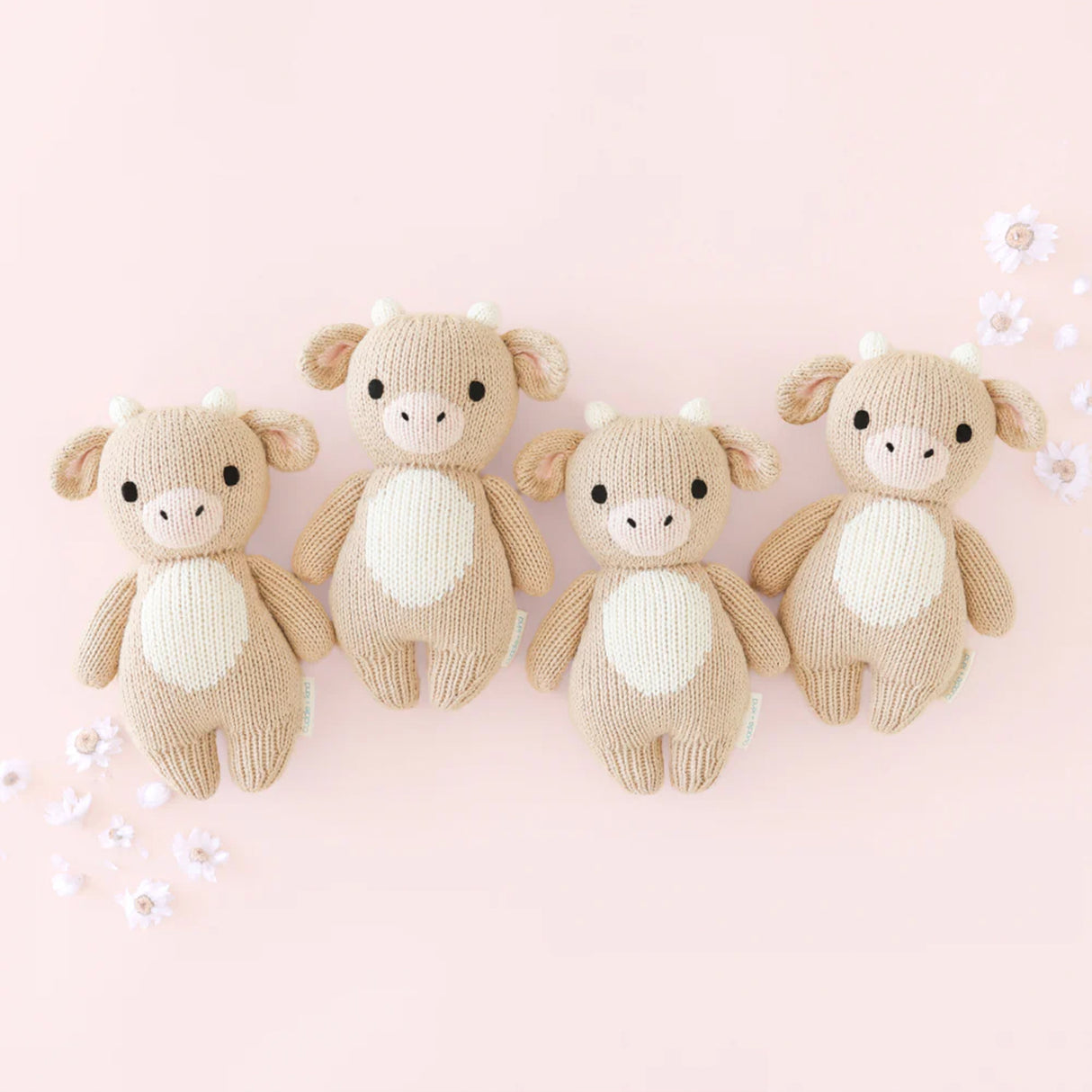 Four knitted baby jersey cow toys on a pink background with white flowers