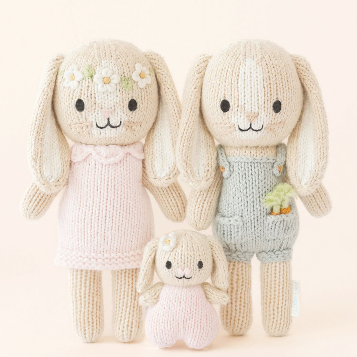 Three knitted bunny dolls one in a pink dress with a flower chain on her head. Next to her bunny toy in a blue dunagree outfit with carrots in his pocket and a baby bunny in between them in pink