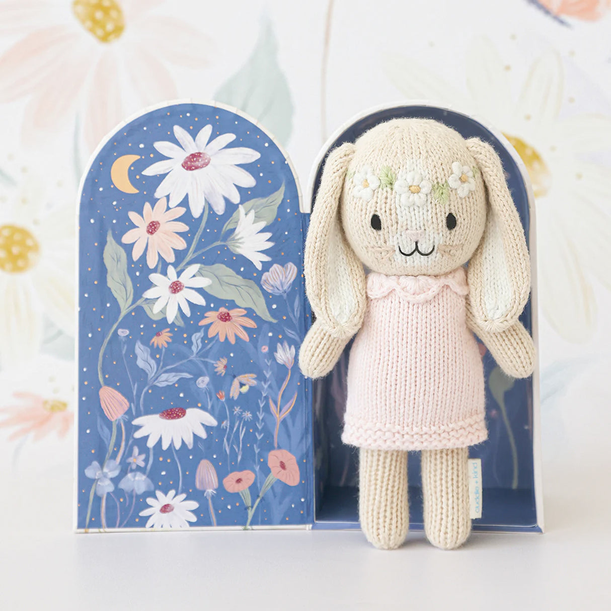 Hand Knitted bunny toy with a pink dress in front of a floral-patterned box on a white background