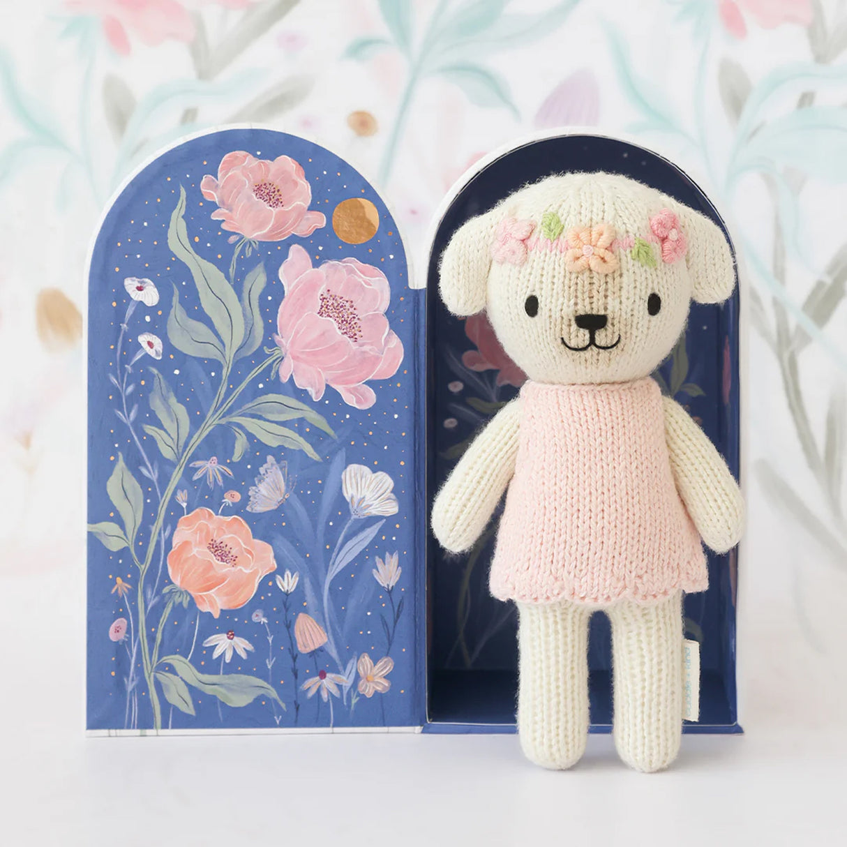 Knitted dog toy in a pink dress with floral decorations, standing next to a decorative box with floral design.