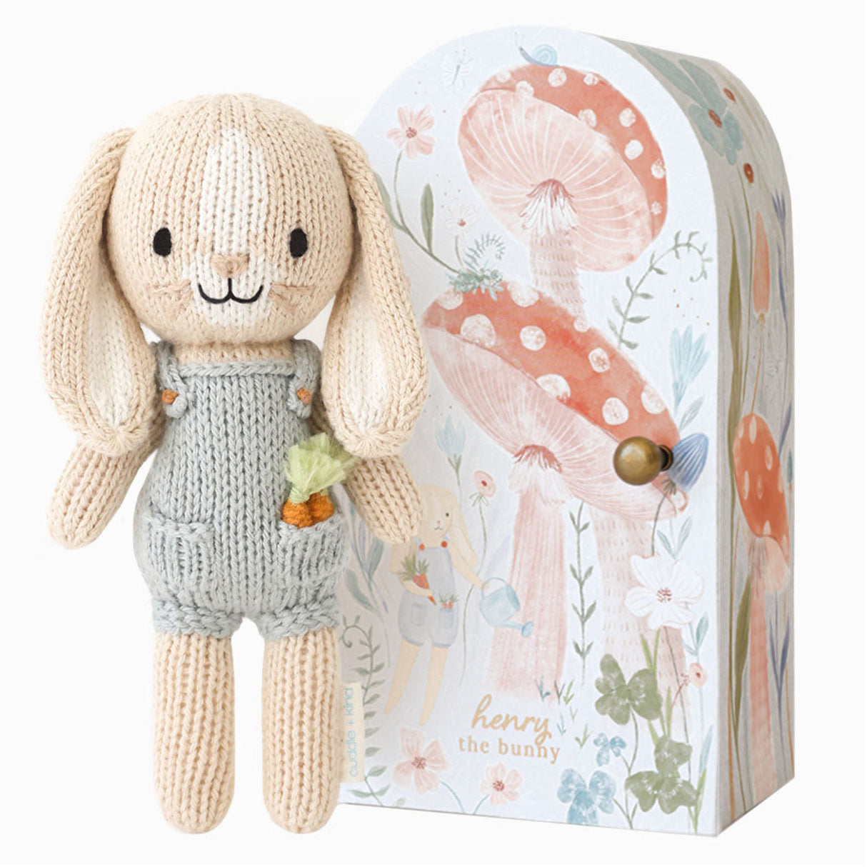 nitted bunny toy in a blue dunagree outfit with carrots in his pocket with a decorative box featuring floral and mushroom designs.