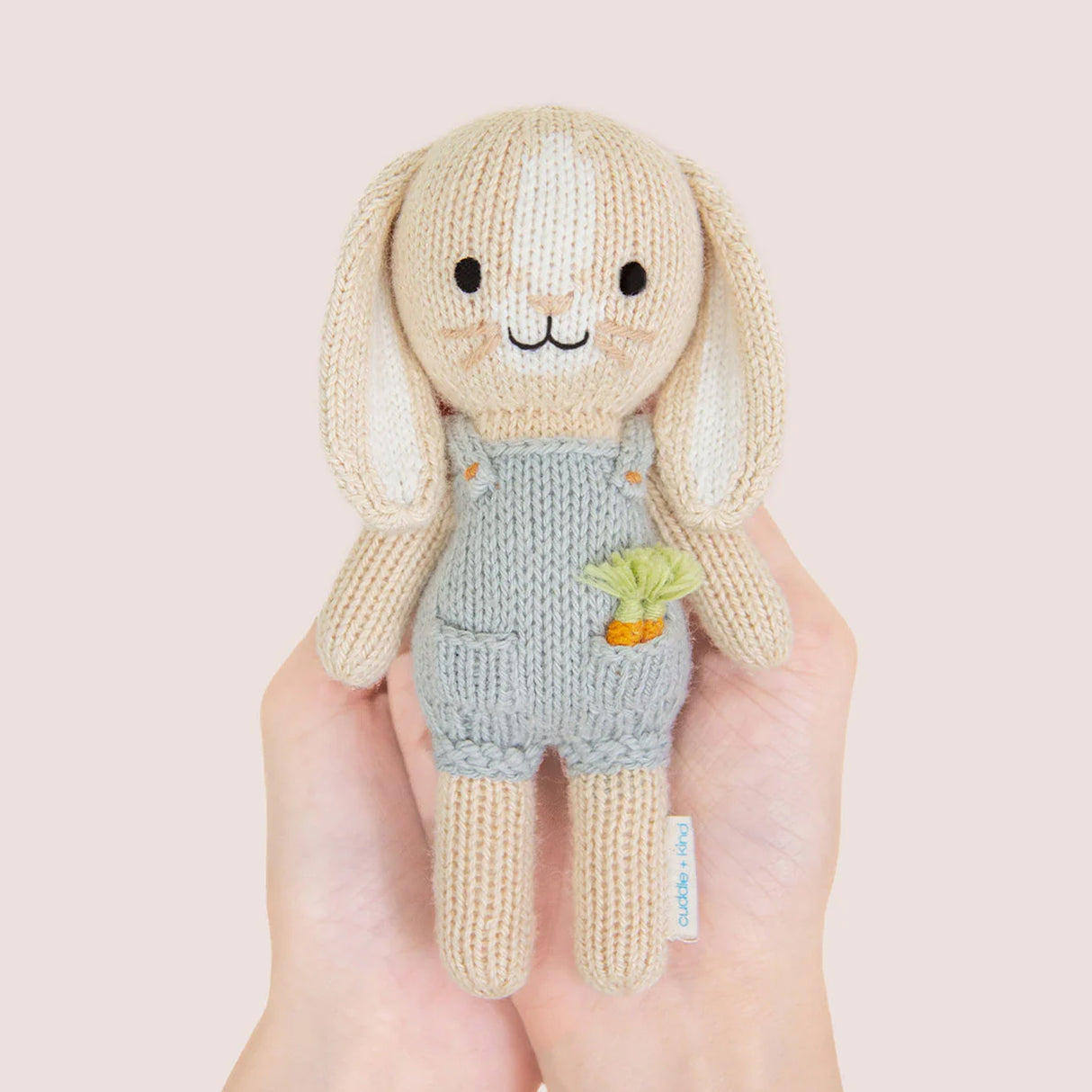 nitted bunny toy in a blue dunagree outfit with carrots in his pocket held by hands against a light pink background
