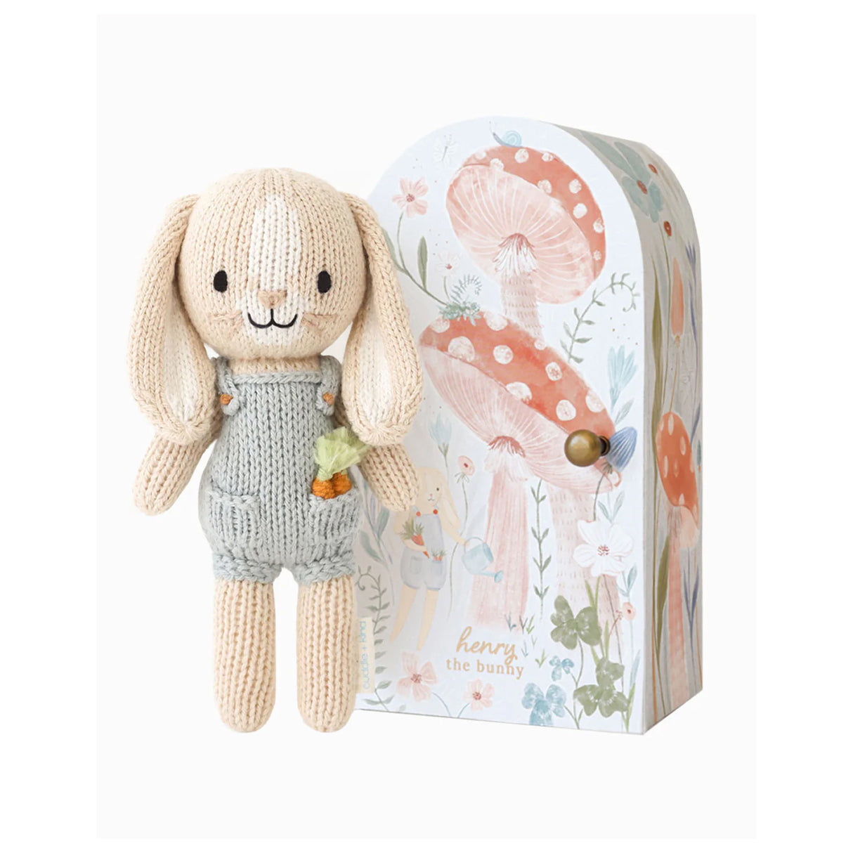Knitted bunny toy in a blue dunagree outfit with carrots in his pocket in front of a decorative box with watercoloured red and white Mushrooms on a white background