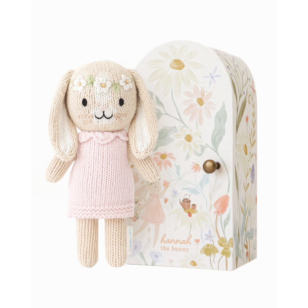 Hand Knitted bunny toy with a pink dress in front of a floral-patterned box on a white background