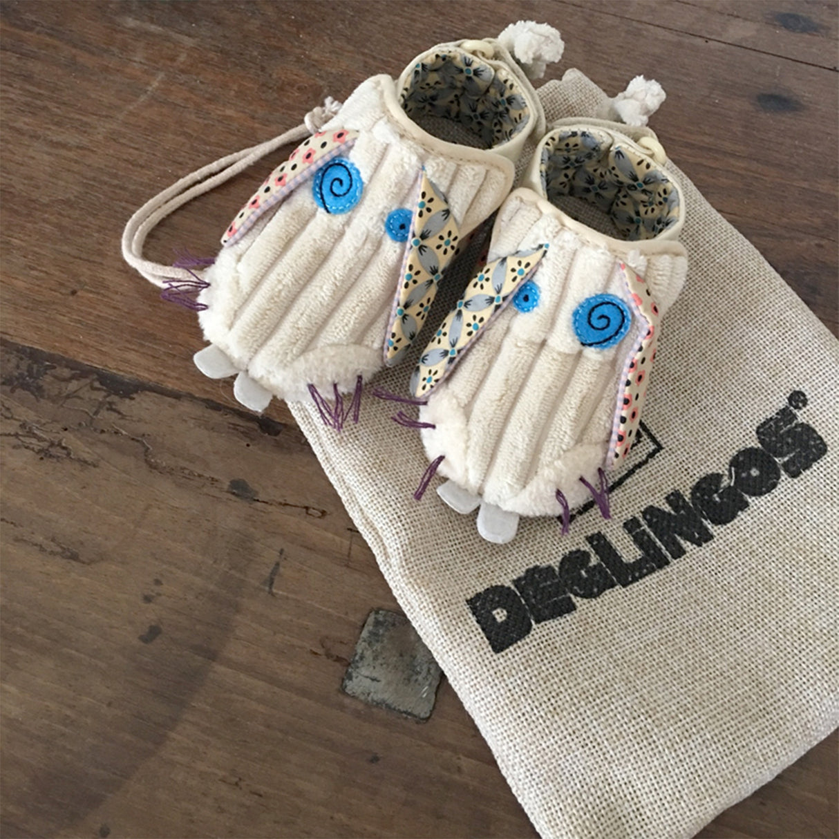 A pair of beige baby booties with blue and purple details, designed to look like a bunny face and tail, with adjustable velcro fasteners.