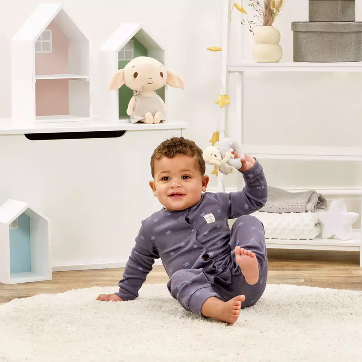 Baby in a blue outfit sitting on a white rug in a playroom with toys and shelves holding a Plush Dobby toy with a ring