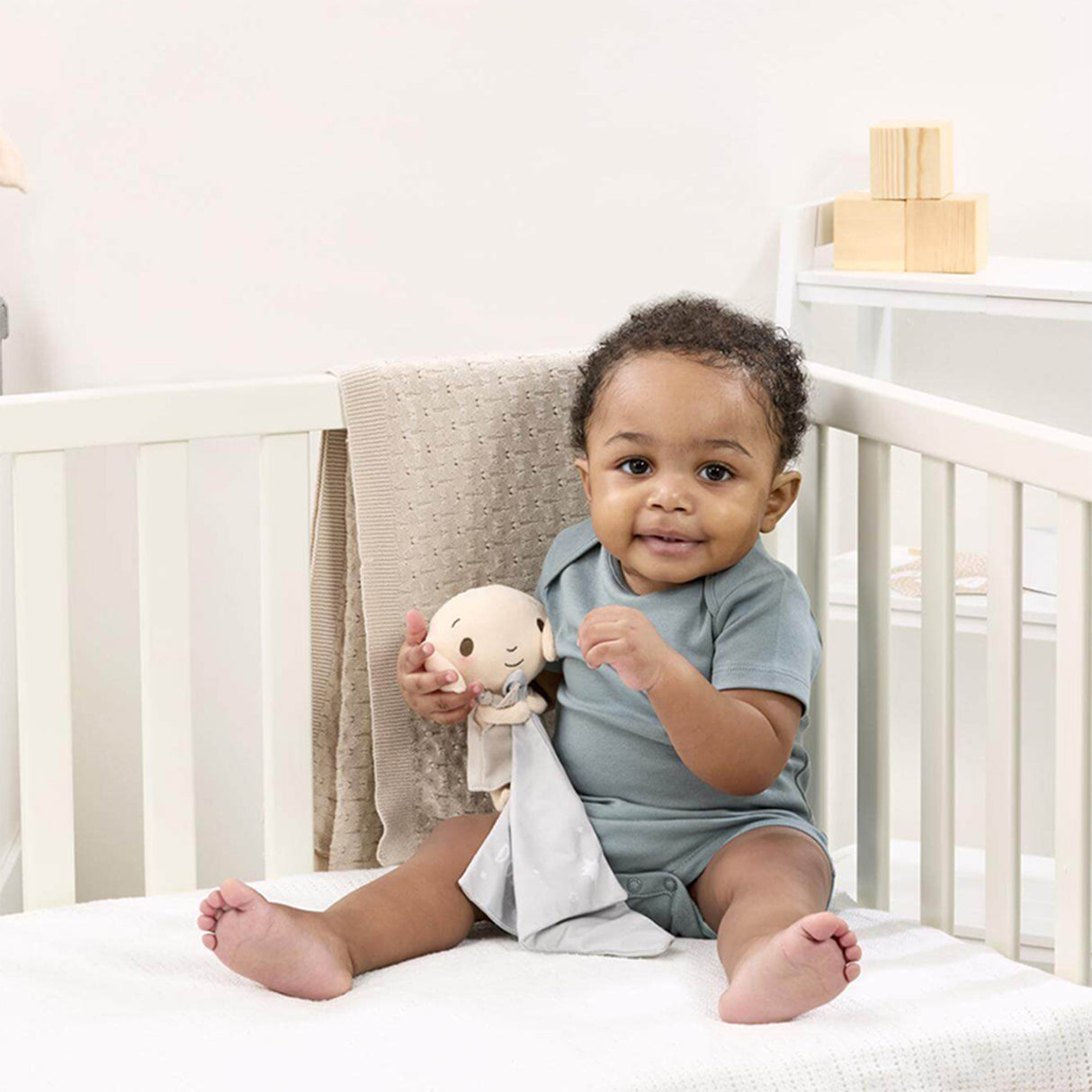 Baby sitting in a crib holding a Harry Potter themed Dobby comforter, featuring a plush Dobby the house-elf on a grey velvety blanket, with a neutral color palette.