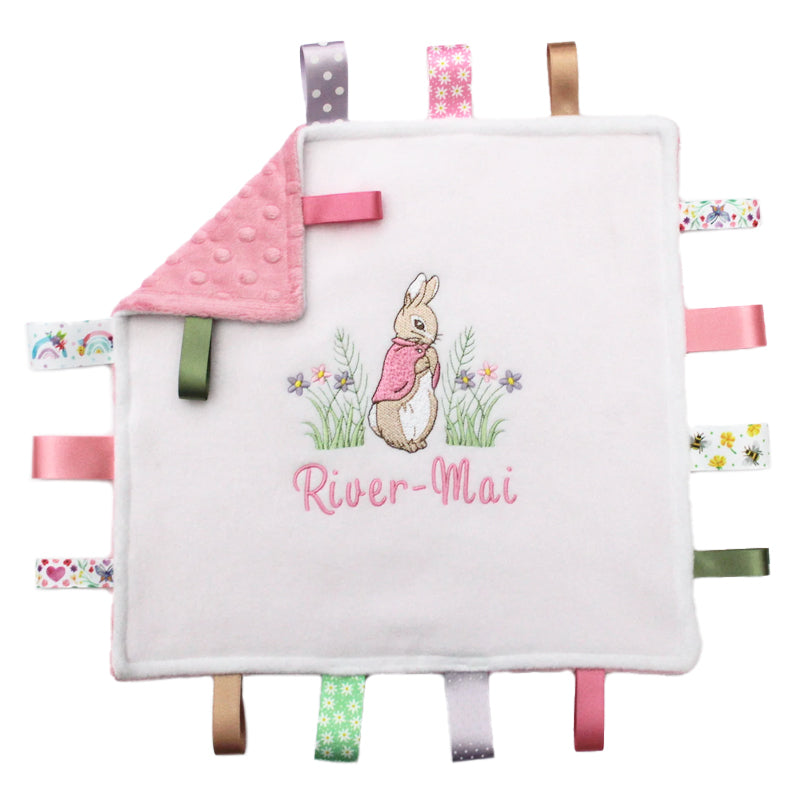 A personalised ribbon taggy blanket with a Flopsy bunny design, featuring a white or cream background with pink / green ribbon tags. The blanket has a Dimple Minky back in pink, white, cream green or and includes decorative satin tags with a variety of patterns.