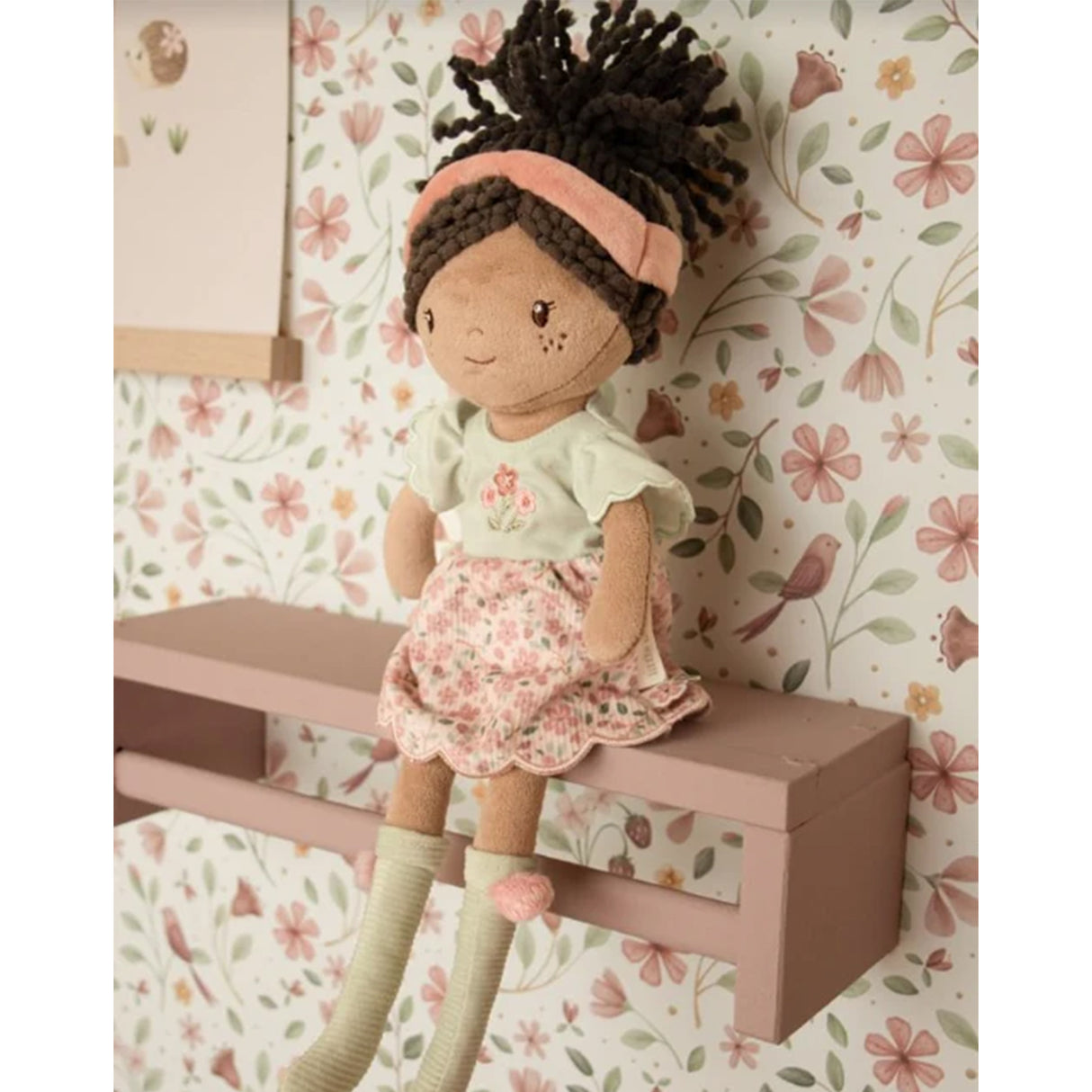 Soft doll with floral pink and sage green dress, pink headband and curly brown hair tied in a ponytail against a floral bird wallpapered background