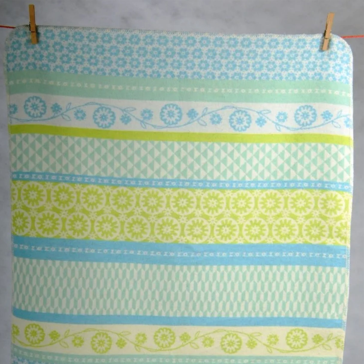 A cotton blanket with retro floral and geometric pattern in light blue and lime green, hanging from two wooden pegs.