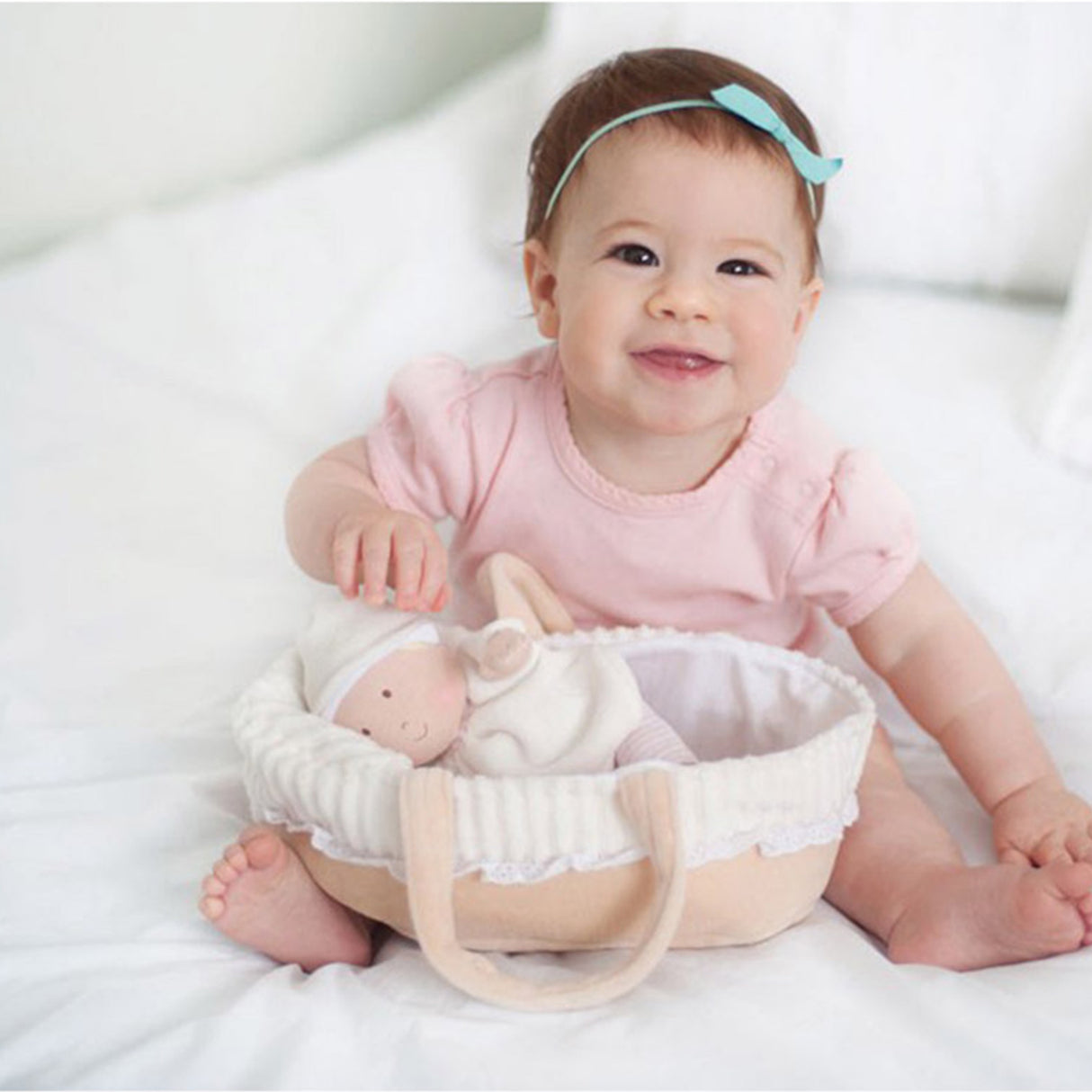 Baby in pink outfit sitting next to a baby doll in a basket on a white background