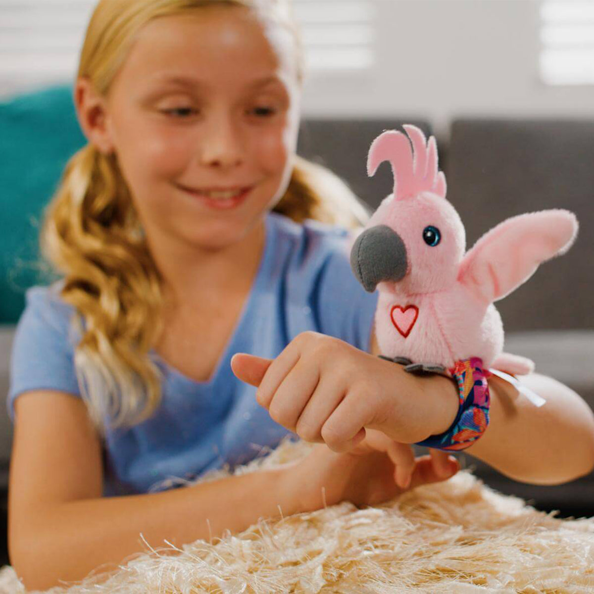 A young girl wearing a colourful wrist strap that holds a pink plush cockatoo toy. The toy has a gray beak, black eyes, and a small red heart on its chest.