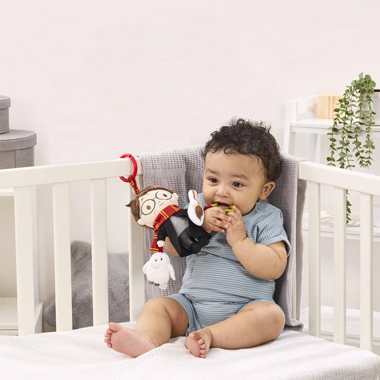 Baby sitting in a crib with a plush Harry Potter toy with round glasses, a lightning bolt scar, wearing Hogwarts robes and a Gryffindor scarf, holding a Firebolt broomstick and accompanied by a squeaking owl toy.