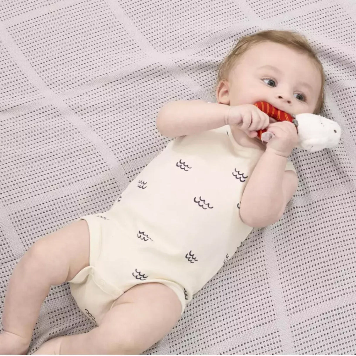 Baby lying on a textured blanket holding a A plush Harry Potter Hedwig baby ring rattle rattle with a white owl on top sitting on a red and orange striped ring.