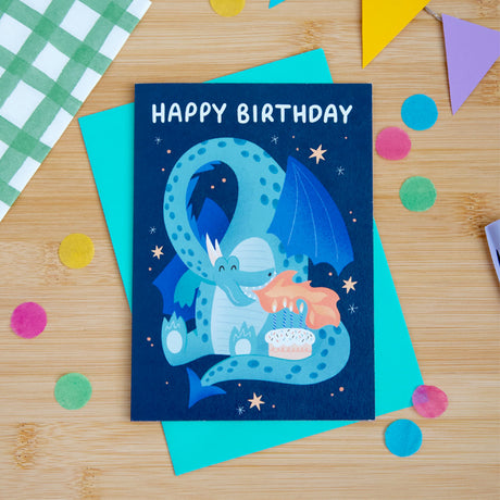 Birthday card with a blue dragon illustration on a wooden surface with colorful confetti.