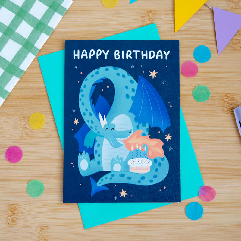 Birthday card with a blue dragon illustration on a wooden surface with colorful confetti.