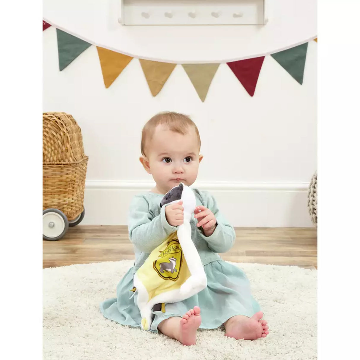 Baby girl sitting holding a Harry Potter themed Hufflepuff comfort blanket with a badger's head, featuring the Hufflepuff crest with yellow and white colours and embroidered stars.
