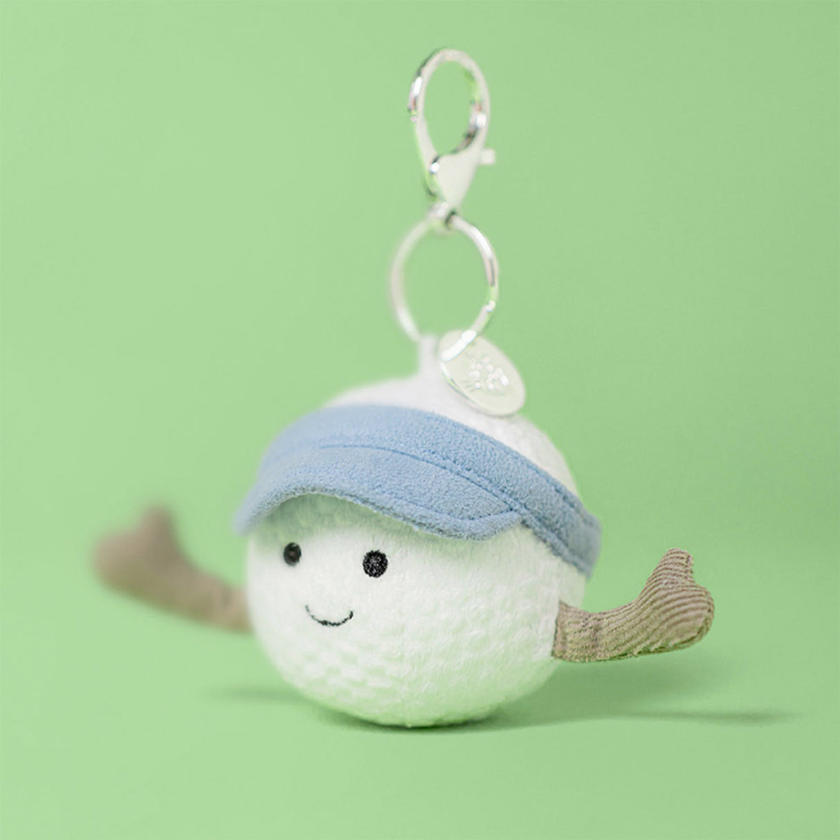 Plush Jellycat golf ball keychain with a blue sun hat and brown arms on a green background