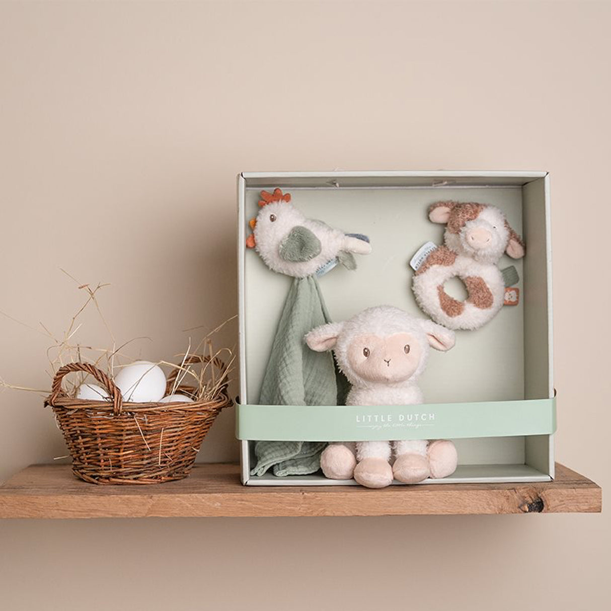 Set of Little Dutch soft toys including a Chicken soother, cow ring rattle, and sheep toy in a Little Dutch sage green box packaging on a wooden shelf with a basket of eggs.