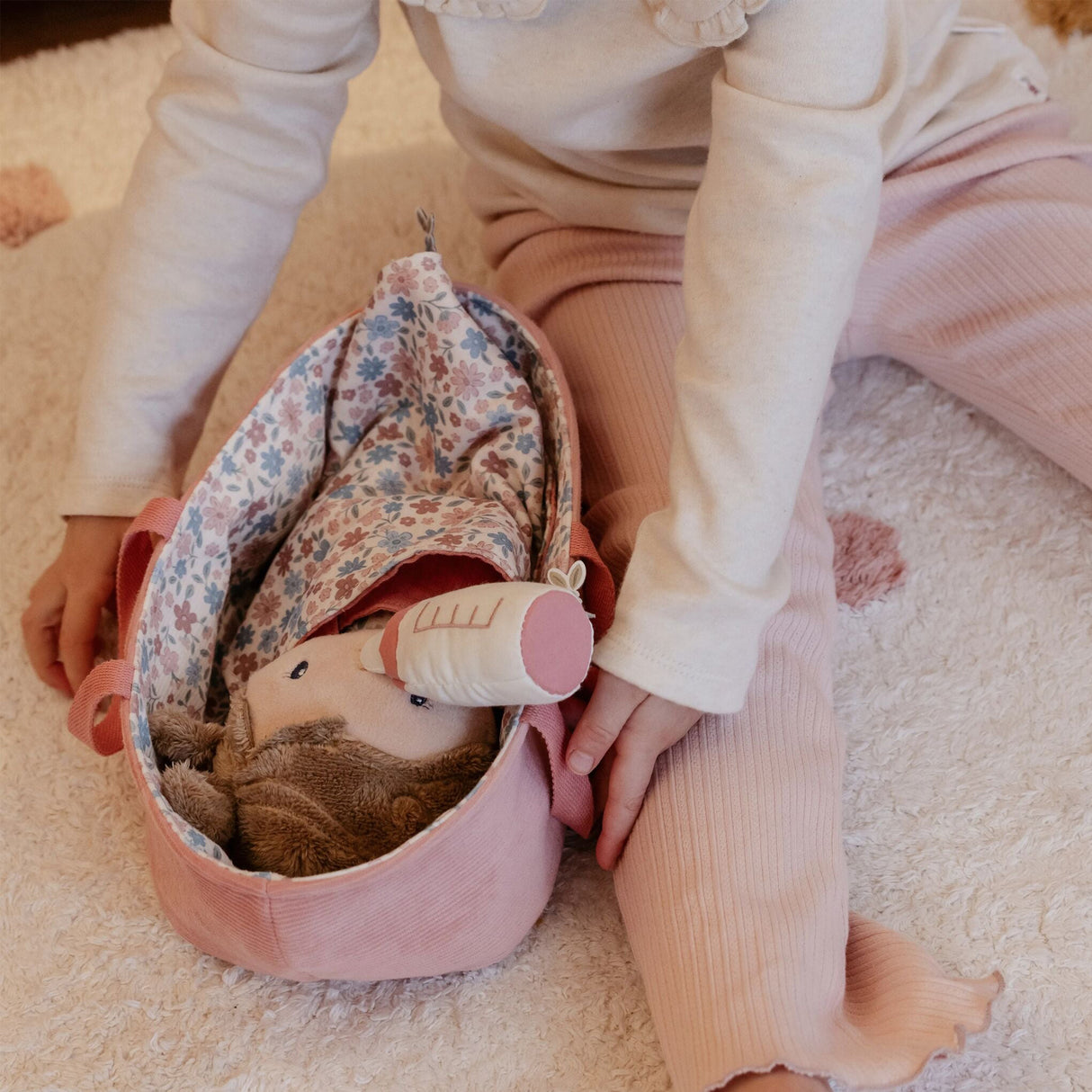 Child holding a pink floral toy carry cot with a soft doll inside on a soft surface