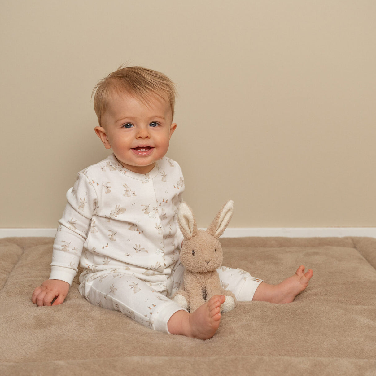 Baby sitting on a beige blanket with a plush toy, smiling at the camera.