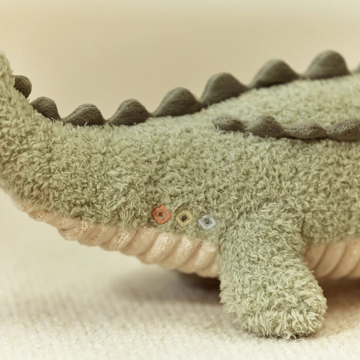 Close-up of a plush crocodile toy made of textured, fuzzy fabric, showing its side with dark green spikes along the back, a cream-colored underside.