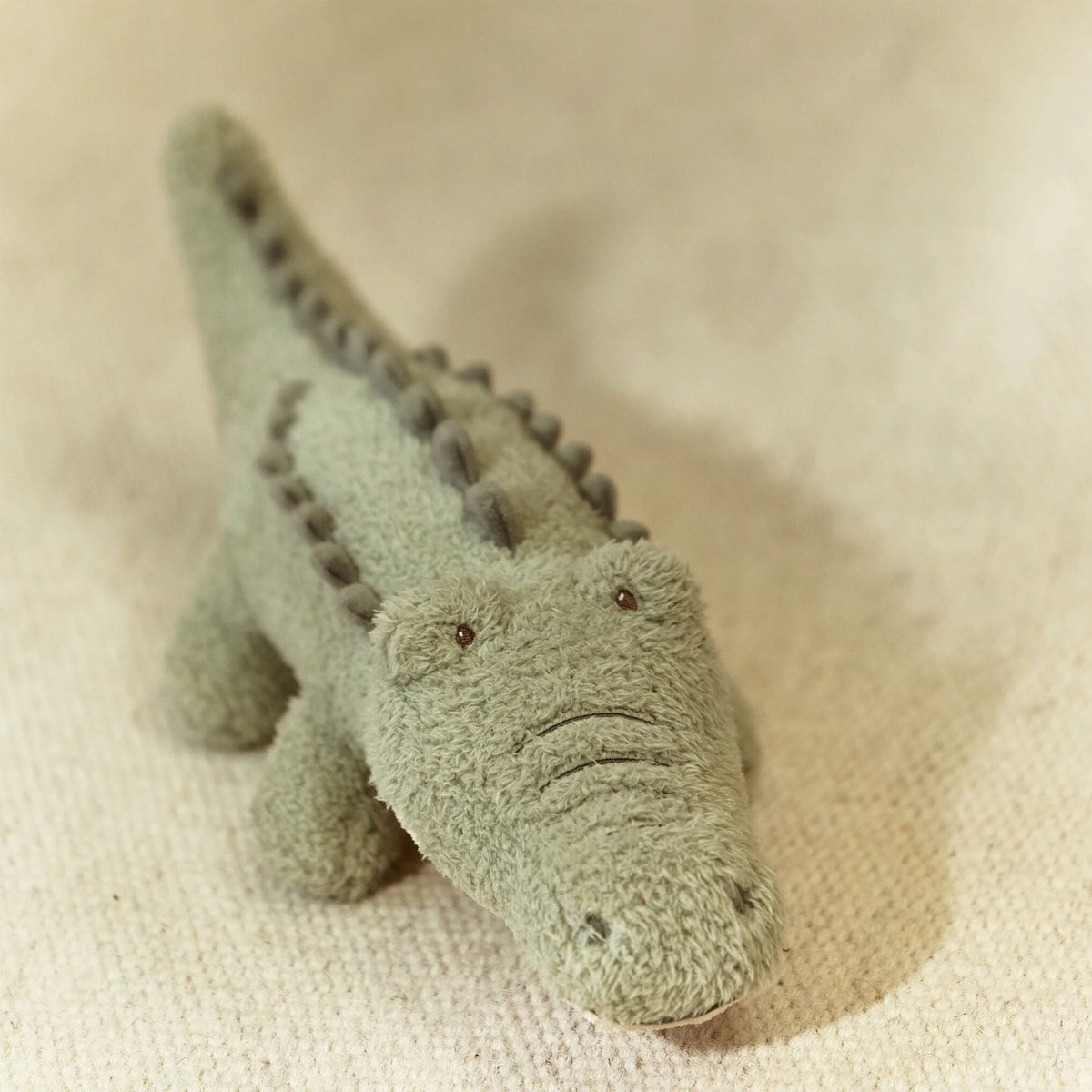 A small plush crocodile toy in a muted sage green color, photographed on a textured beige fabric background.