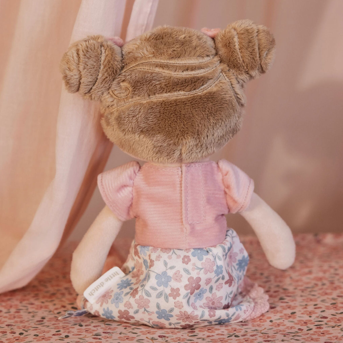 Back view of the Little Dutch Hug Doll Rosa sitting, showing brown fluffy bun hairstyle, pink T-shirt and floral skirt with Little Dutch label