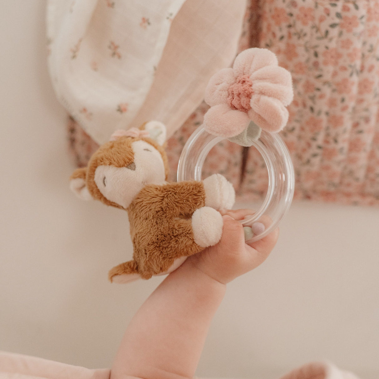 Child's hand holding a plush toy with a clear ring and pink flowers, against a floral background.