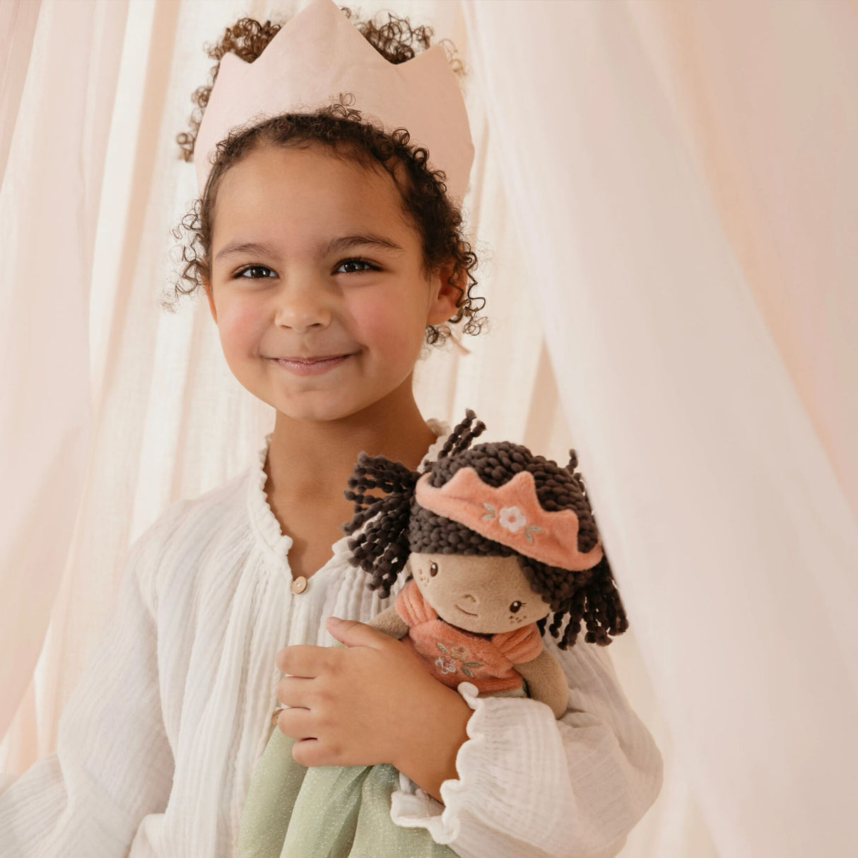 A young child wearing a light pink crown and white textured blouse, holding a small doll with dark curly hair