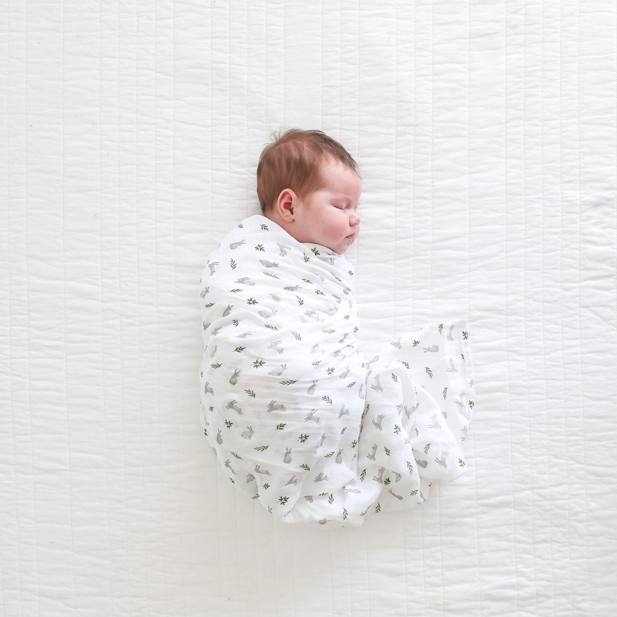 Newborn baby swaddled in a white blanket with bunny and leaf pattern on a textured white surface