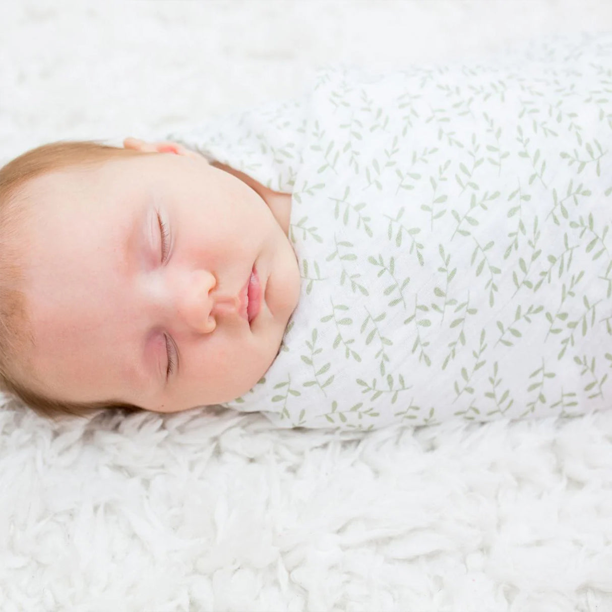 Newborn baby sleeping peacefully wrapped in a white leaf patterened swaddle.
