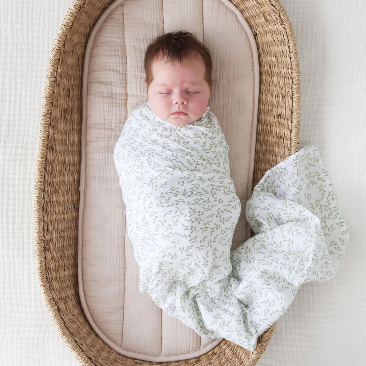 Newborn baby wrapped in a leaf patterened swaddle blanket lying in a wicker crib.