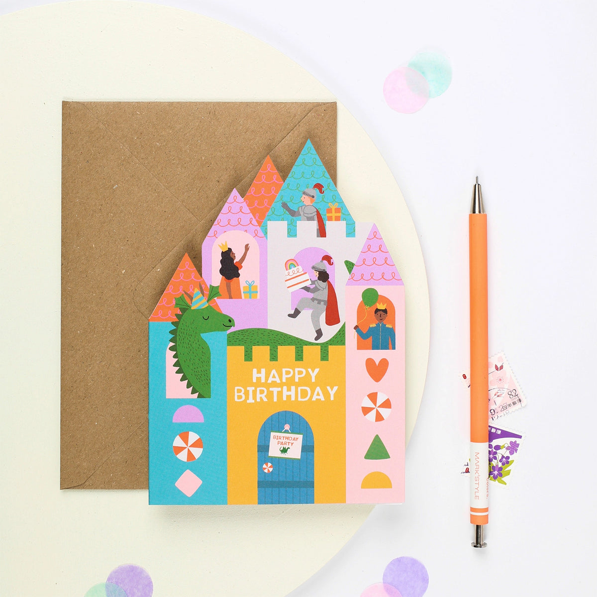 Mifkins Fairytale Castle birthday card with kraft envelope next to an orange pen, showing colourful castle design with dragon and fairytale characters