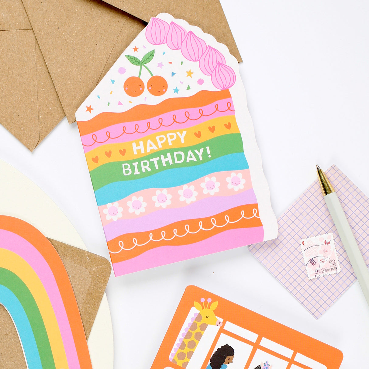 Mifkins Colorful birthday card with cake design on a white surface with stationery items.